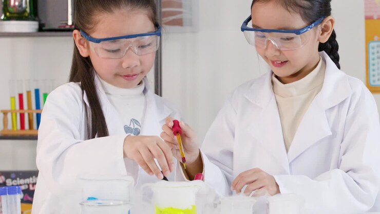 Two young girls wear lab coats and safety goggles while mixing colorful chemicals in beakers
