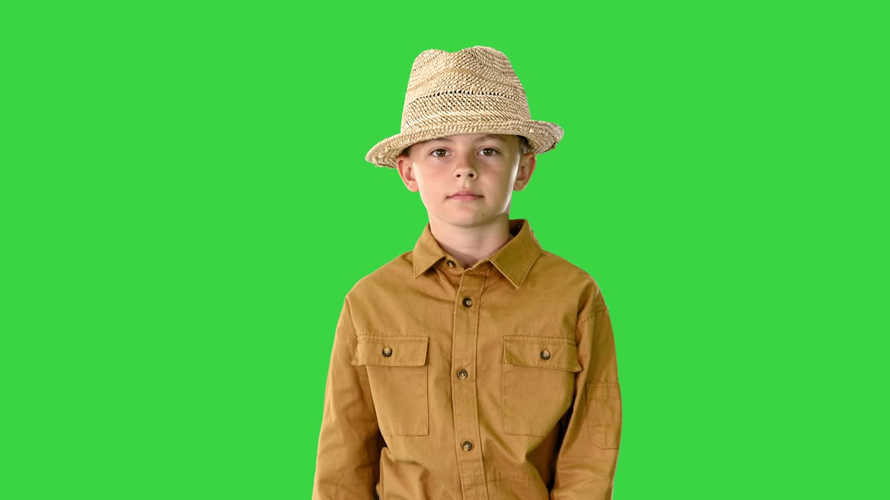 niño con una camisa ajustando su sombrero de paja mientras camina y mira a la cámara en una pantalla verde, clave de croma