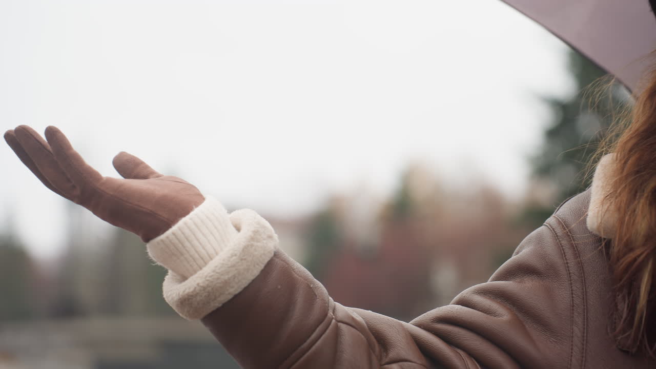 Close-up shot of female standing under light snowfall, holding an umbrella and stretching her hand out. She wears a black knit cap and brown shearling jacket, enjoying peaceful winter atmosphere