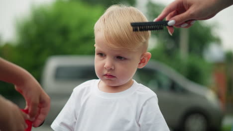 a young boy stares thoughtfully while a woman with painted nails gently combs his hair, the boy seems lost in thought, observing someone near him closely, a car is seen behind them