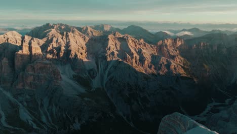 Scenic-aerial-drone-view-of-the-rugged-Dolomite-mountains-in-Italy-at-sunrise,-with-a-snowy-foreground-slope-leading-to-the-peaks
