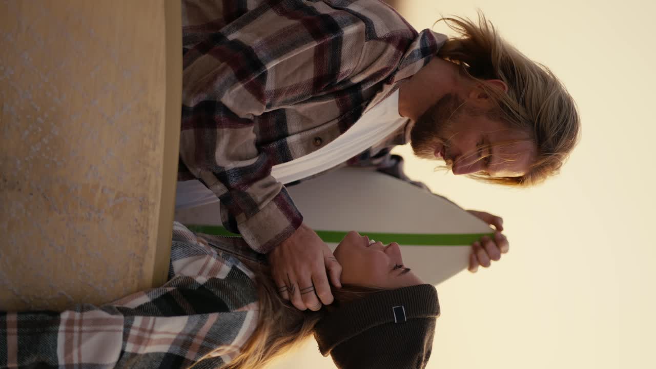 video vertical: un chico rubio con cabello largo y barba en una camisa a cuadros se mantiene cerca y se comunica con su novia rubia en una camiseta a cuadros que lo mira y le toca el cabello con la mano mientras sostiene una tabla de surf en la mano por la mañana cerca del mar