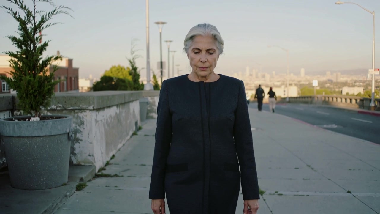 Walking senior woman wearing dark coat starting toward camera on urban sidewalk, with concrete wall