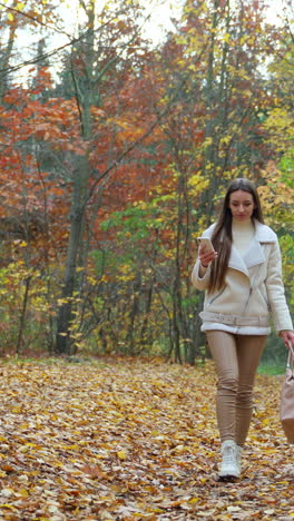 attractive romantic girl with bag and phone in her hands walks by the park. calm and peaceful time spending in the autumn nature. vertical video