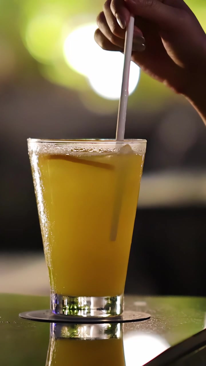 Mano mescola un cocktail giallo con cannuccia in un bicchiere, illuminazione bokeh serale, primo piano.