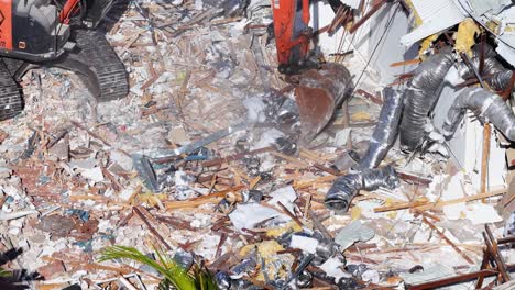 an excavator uses its bucket to demolish a house, scattering debris and dust in a sunlit construction site on the gold coast, australia