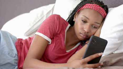 african american young woman lying on bed, looking at smartphone