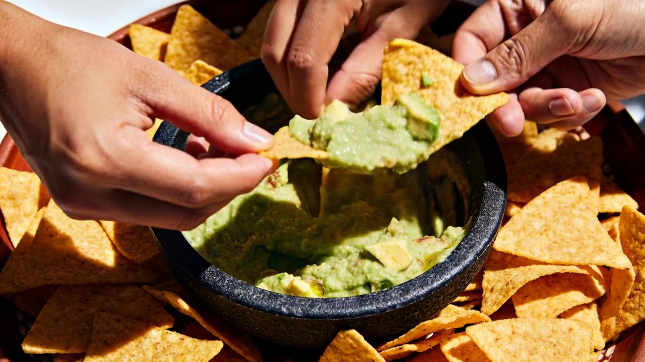 Dvě dospělé ruce sahají po svačině, namáčejí a nabírají guacamole v kuchyni, s kamennou miskou a chipsy