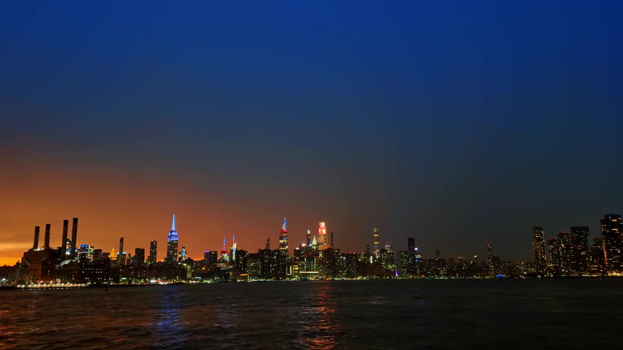 Midtown Manhattans bybillede om natten over vandet. Oplyste skyskrabere spejler sig i floden i den mørkeblå aftentime.