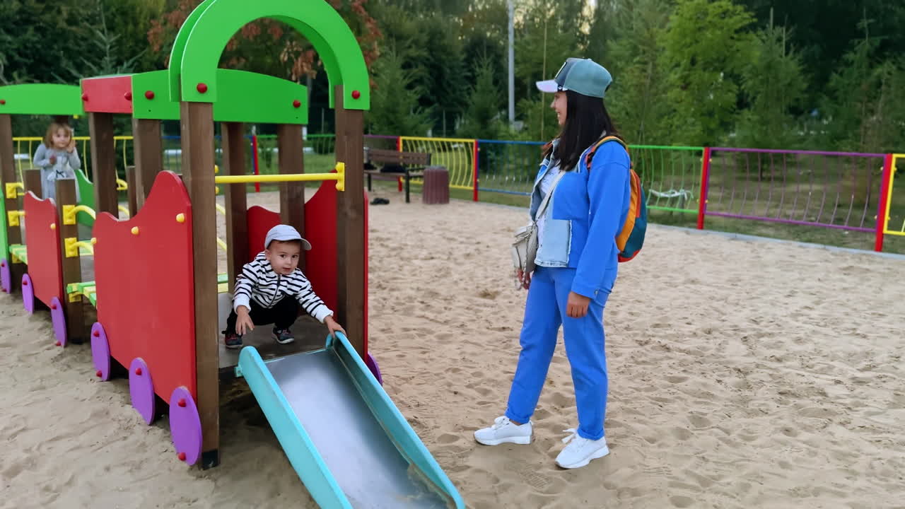 Cute baby boy stands in the toy train at playground. Mother stands watching beside. Kid on the walk.