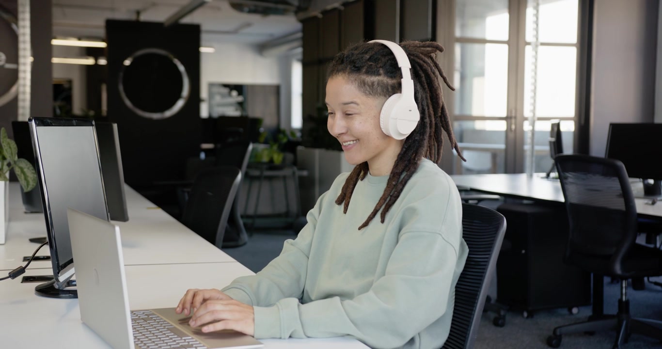 Nonbinary worker hearing through headphones, working on laptop at work, smiling at desk, copy space