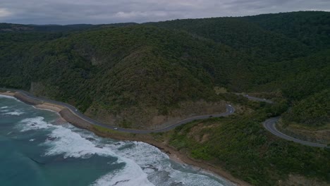 Tilt-drone-aerial-view-of-famous-Great-Ocean-Road-coastal-highway-with-car-traffic-and-forest-mountains,-Victoria,-Australia