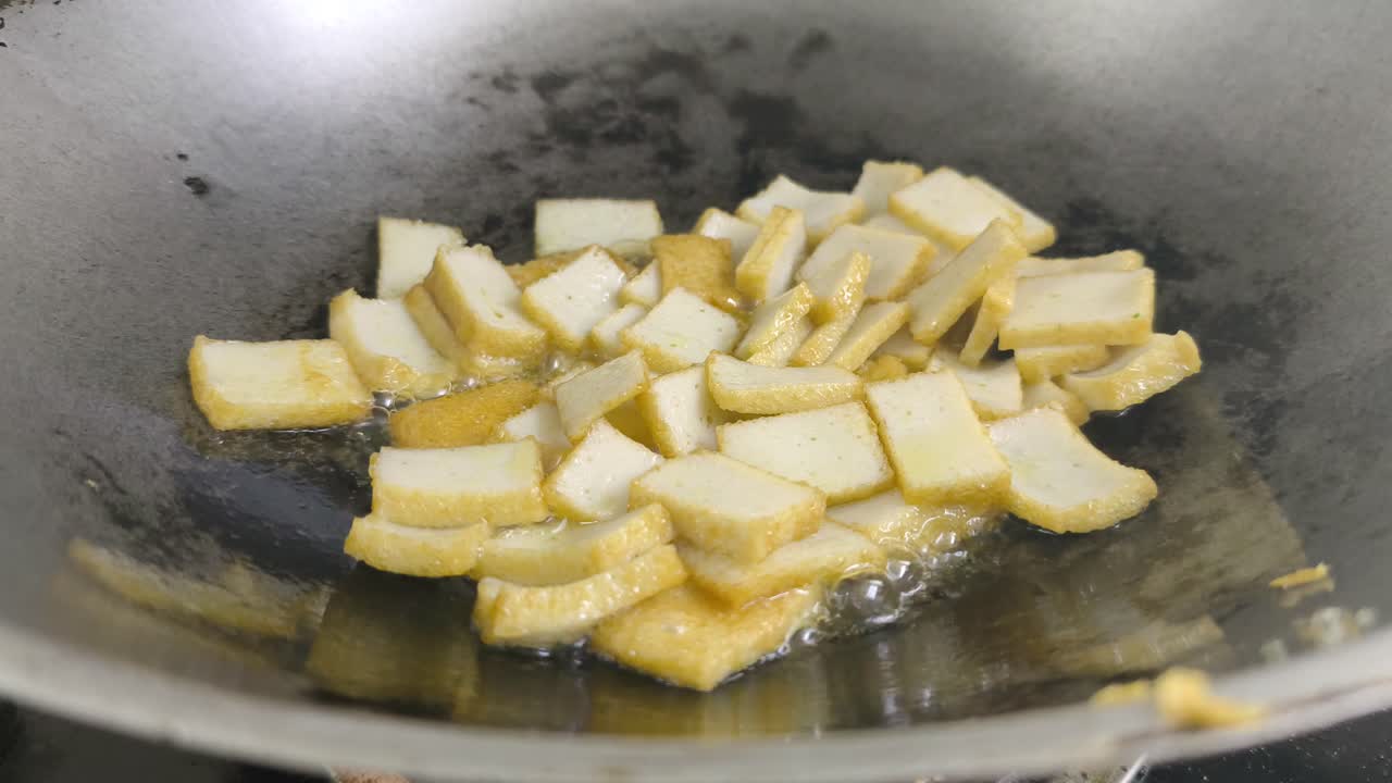 Chef frying sliced fish cake in a pan using palm oil. The chef uses a spatula to turn the pieces, ensuring they cook evenly