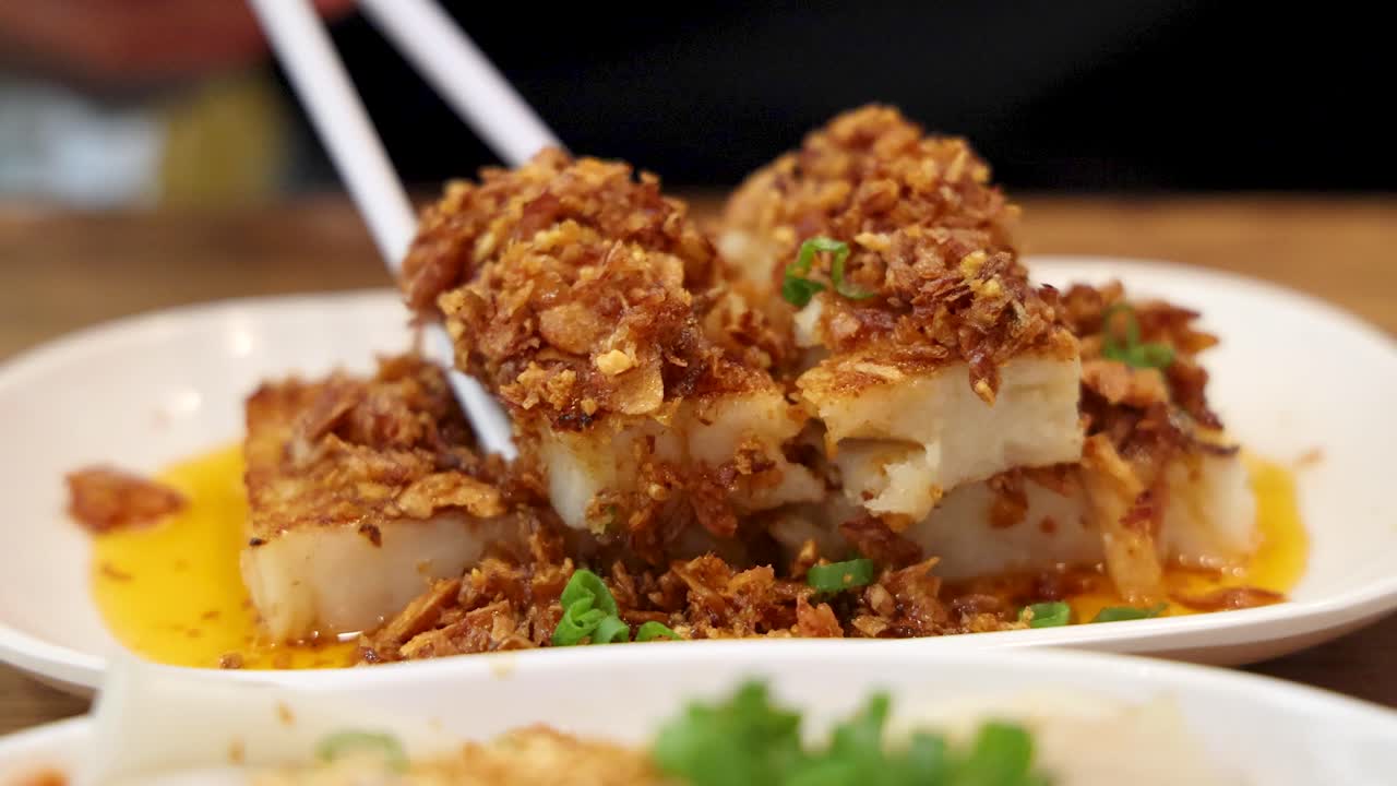 Chopsticks pick up a piece of fried turnip cake topped with crispy shallots and herbs, under warm indoor lighting, close-up perspective, shallow depth of field