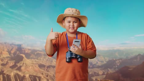 asian boy with a hat and binoculars smiling and pointing to smartphone while traveling at the top of mountain. boy researcher examines something, travel tourism adventure concept