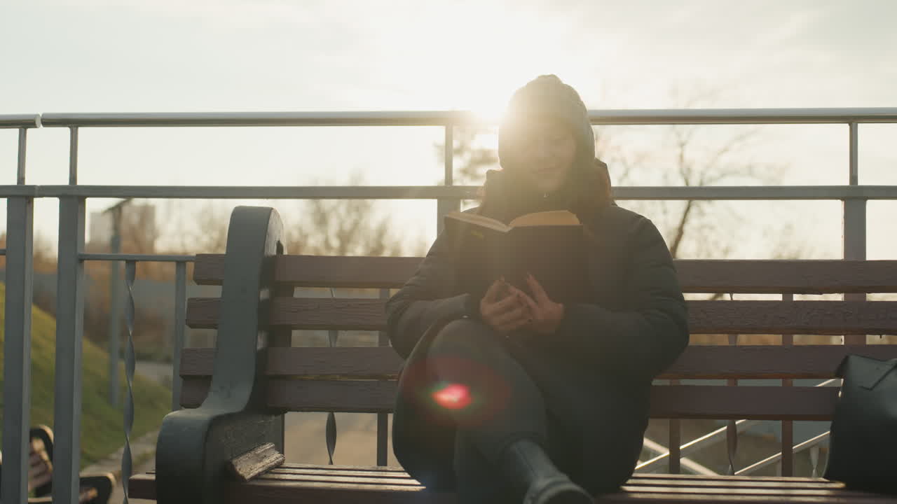 Young woman reading at sunset bench with warm halo and distant hills. Travelerstyle break on urban walkway, backpack nearby, beanie and padded coat, gentle smile as she turns pages, soft lens flare