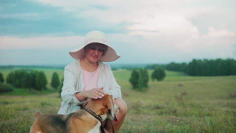 dog owner in sunhat squatting near her pet, holding its leash while gently rubbing its back, the background features rolling green fields and trees