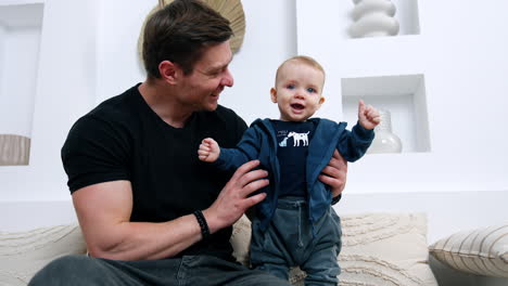 father holds his lovely smiling baby boy standing beside on the sofa. dad and son are at home.