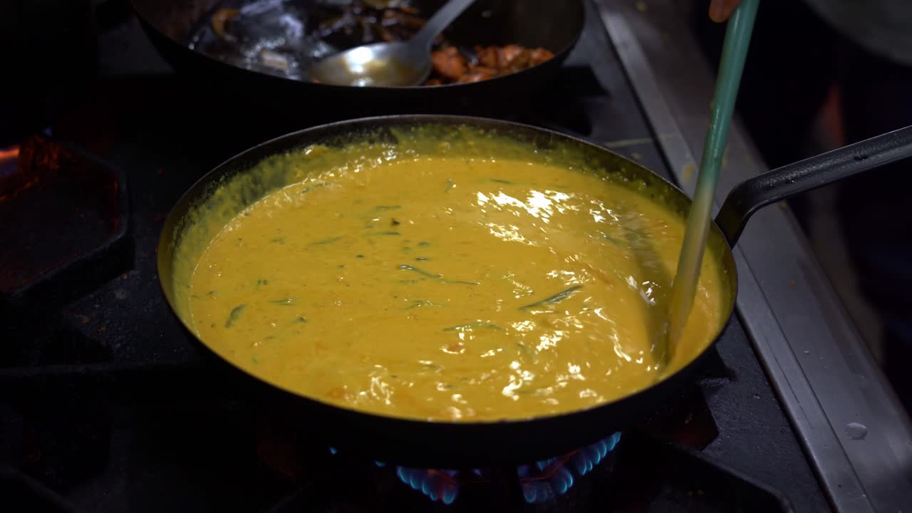 chef usando una espátula revolviendo el delicioso curry amarillo con hojas frescas de curry en una sartén hirviendo caliente, cocinando cocina india tradicional en un ambiente de cocina comercial