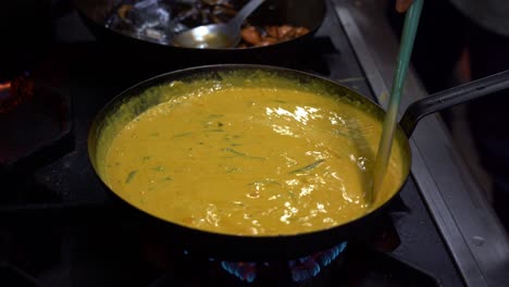 Chef-Usando-Una-Espátula-Revolviendo-El-Delicioso-Curry-Amarillo-Con-Hojas-Frescas-De-Curry-En-Una-Sartén-Hirviendo-Caliente,-Cocinando-Cocina-India-Tradicional-En-Un-Ambiente-De-Cocina-Comercial