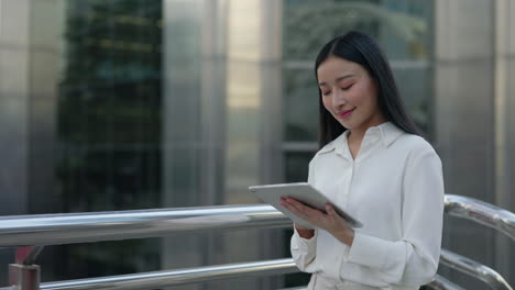 young asian woman smiling while using a tablet outdoors in an urban setting