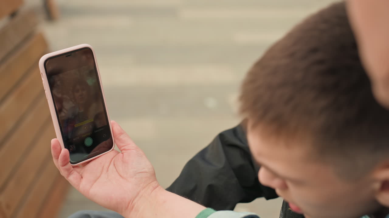 White Teenage Boys Watch Phone Screen During Outdoor Video Call On Bench, Handheld Smartphone Displaying Family Portrait, CloseUp Of Hand And Screen, Spontaneous Laughter And Reactions, Modern