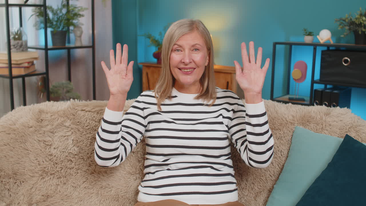 Mature woman waving hello hi on sofa smiling warmly expressing friendliness and openness at home