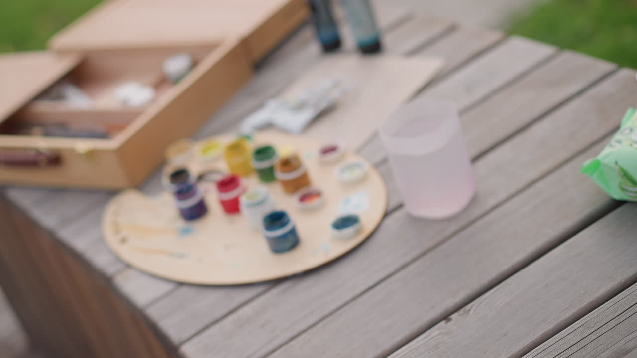 wooden paint box with palette and brushes on outdoor bench, portable plein air painting setup with jars of gouache and acrylic paints, clear water cup for rinsing brushes, open compartments holding