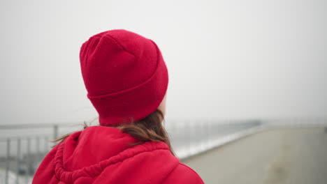primer plano de una mujer con gorra roja y sudadera con capucha corriendo al aire libre durante el invierno cerca de la barandilla de hierro por el puente en una atmósfera de niebla, mostrando fitness y un estilo de vida activo en un entorno urbano sereno