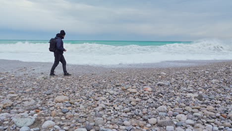 Ein-Mann-Schlendert-über-Einen-Steinigen-Strand,-Während-An-Einem-Trüben,-Bewölkten-Morgen-Tosende-Wellen-Ans-Ufer-Krachen