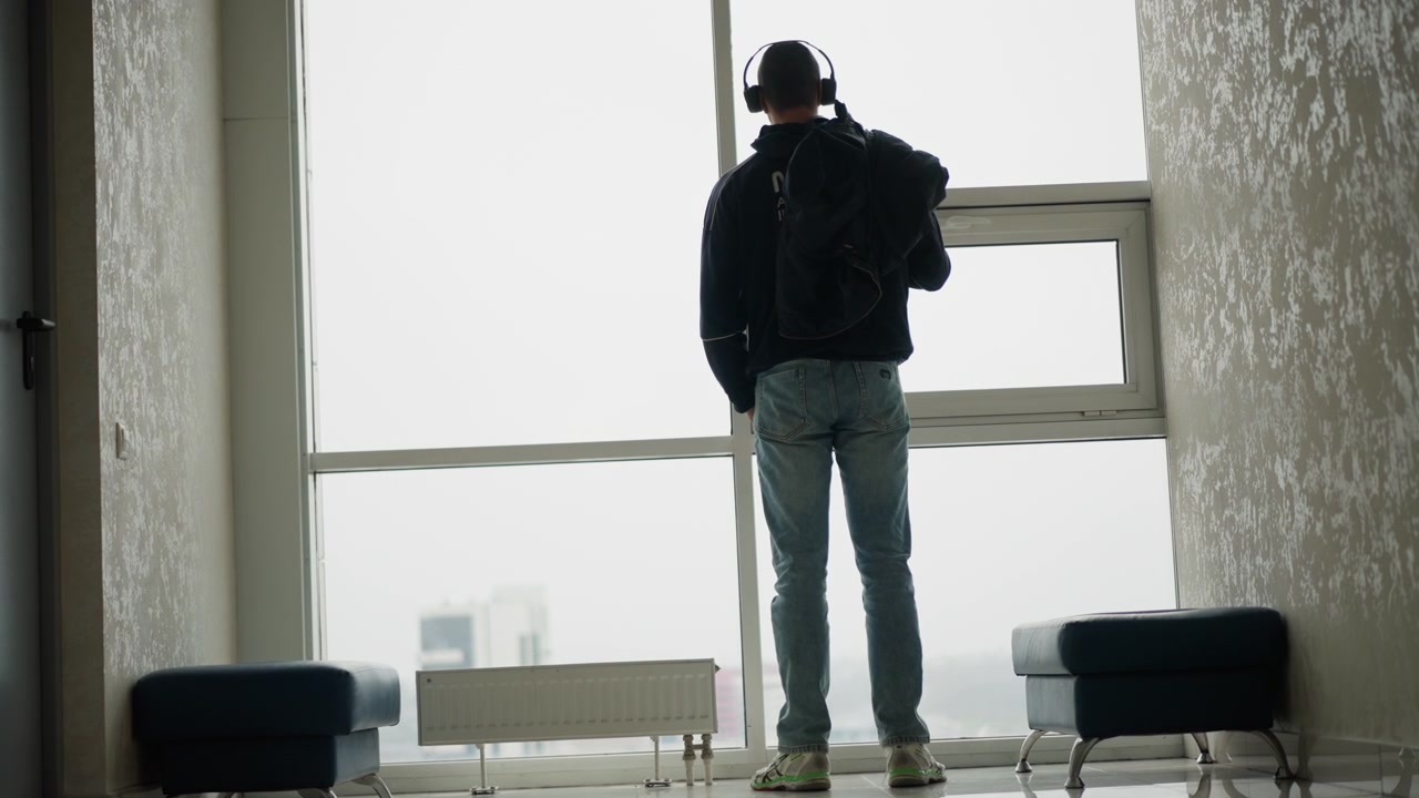 Close up of man wearing headphones and carrying backpack standing still in front of large windows. scene features soft lighting with overcast daylight streaming through window revealing