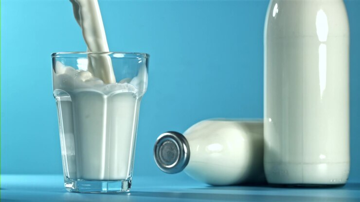 Rallentatore: Versando latte fresco in un bicchiere con bottiglie su sfondo blu.