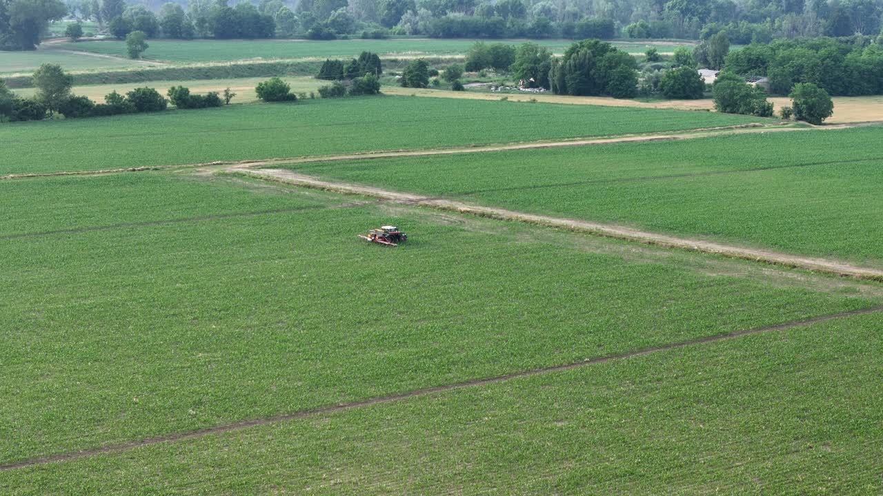 Aerial view of tractor with boom sprayer working across a lush green maize field, applying treatments to support crop health and productivity in a carefully managed rural farming landscape, PC, Italy