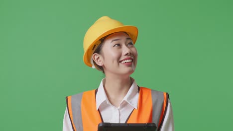 close up of asian female engineer with safety helmet looking at the tablet in her hand and looking around while standing in the green screen background studio