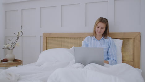 young millennial girl sitting on a bed in the morning uses laptop computer. caucasian woman home urban working online laptop bedroom bed