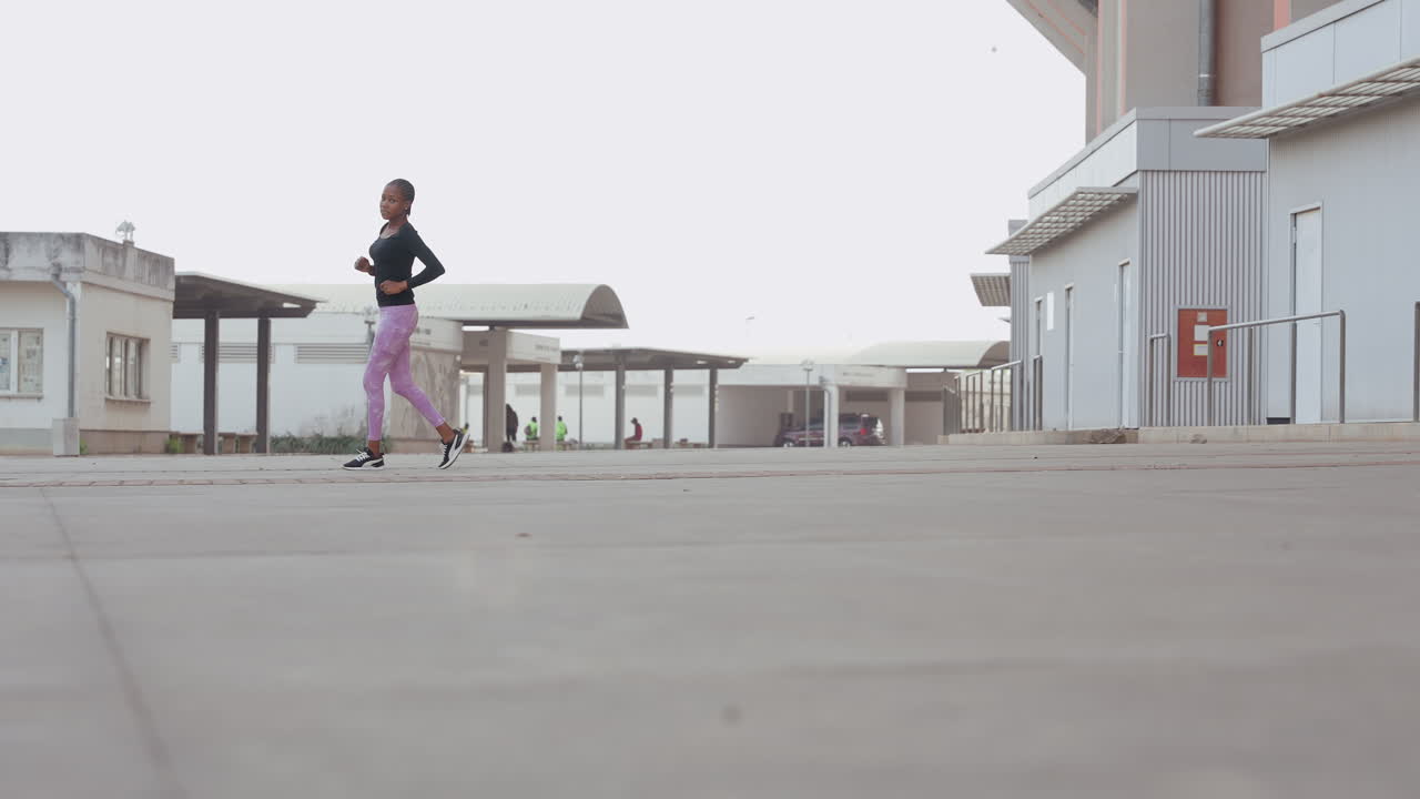 Ampia inquadratura dal basso che mostra una donna correre attraverso una distesa di cemento, una lunga prospettiva enfatizza il terreno e il movimento, una maratoneta in allenamento che avanza in una piazza industriale vuota all'alba con passo costante.