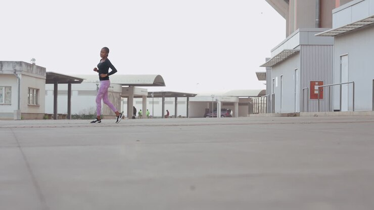 Ampia inquadratura dal basso che mostra una donna correre attraverso una distesa di cemento, una lunga prospettiva enfatizza il terreno e il movimento, una maratoneta in allenamento che avanza in una piazza industriale vuota all'alba con passo costante.