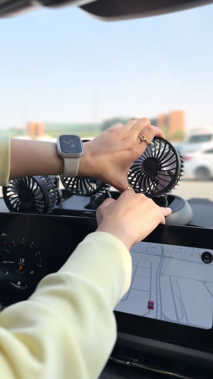 Mani che regolano tre ventilatori portatili di raffreddamento sul cruscotto di un'auto accanto a un display di navigazione, creando un flusso d'aria extra nell'abitacolo di un veicolo parcheggiato in pieno giorno