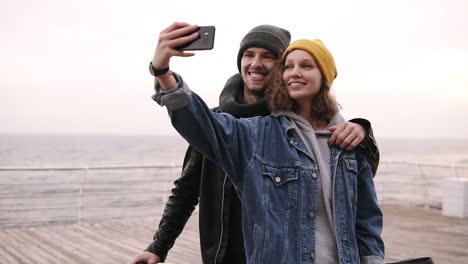 Happy-Young-Couple-Taking-Selfie-By-Mobile-Phone-Near-The-Seaside-In-Cloudy-Day