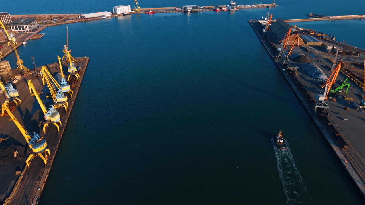 Pemandangan udara kapal tunda yang menavigasi melalui kanal pelabuhan. Tampak atas kapal tunda bergerak di air biru tua di antara dua dermaga industri dengan derek.