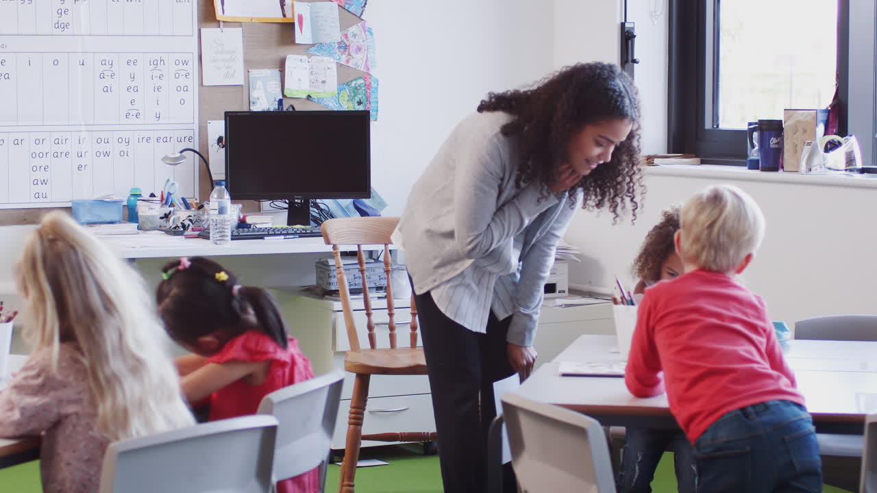 Female infant school teacher leaning at desk helping school kids in a classroom