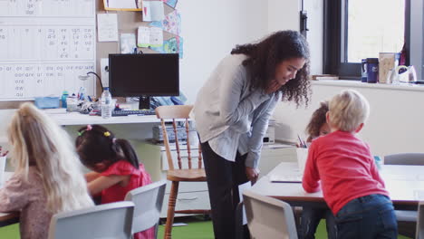 female infant school teacher leaning at desk helping school kids in a classroom