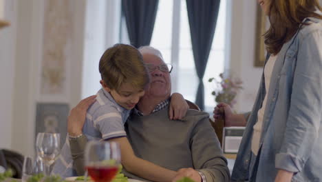 Senior-White-Haired-Man-In-Glasses-Sitting-At-Table-At-Family-Party-While-Talking-To-Granddaughter-And-Grandson-And-Hugging-Them