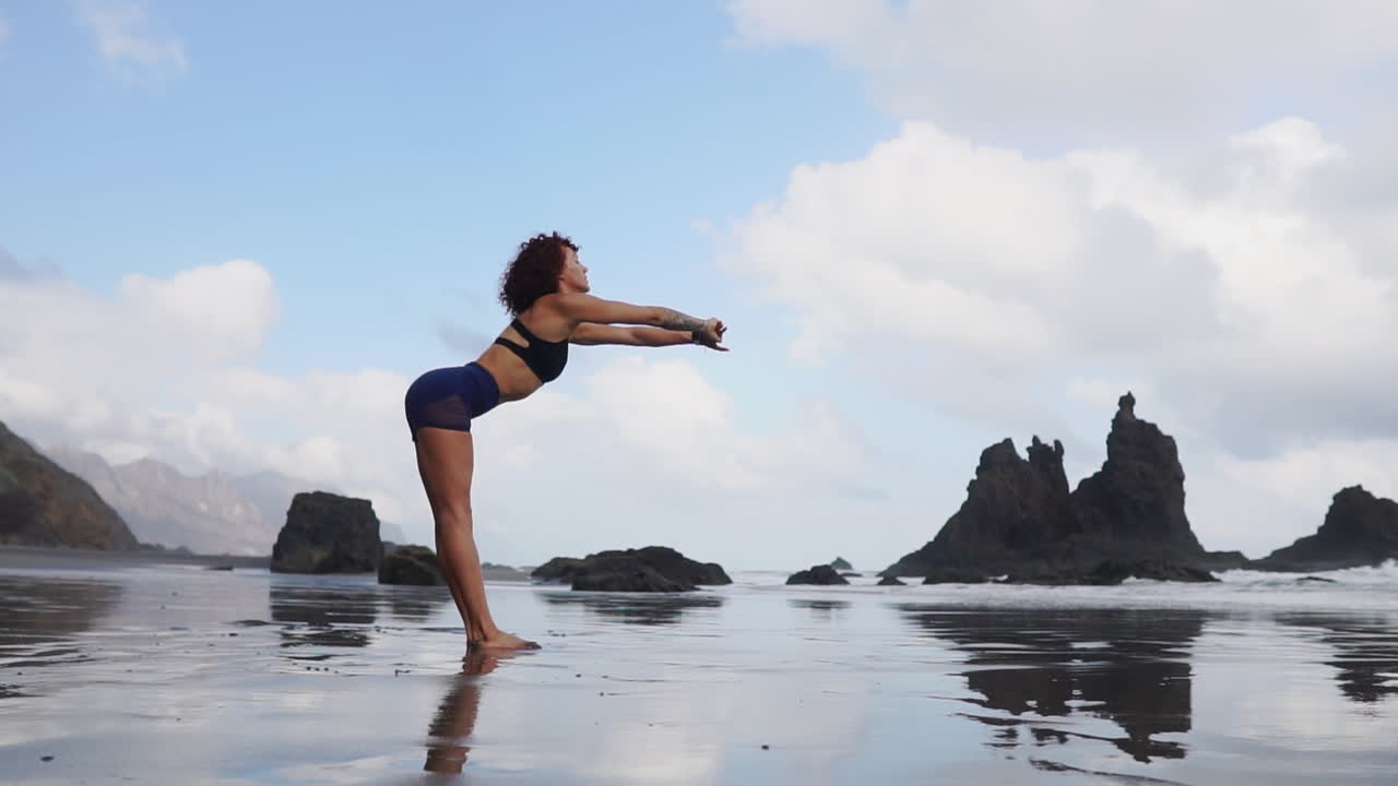 Slow motion reveals a young, slim woman's peaceful practice of stretching and yoga near the ocean, her eyes focused on the distant view