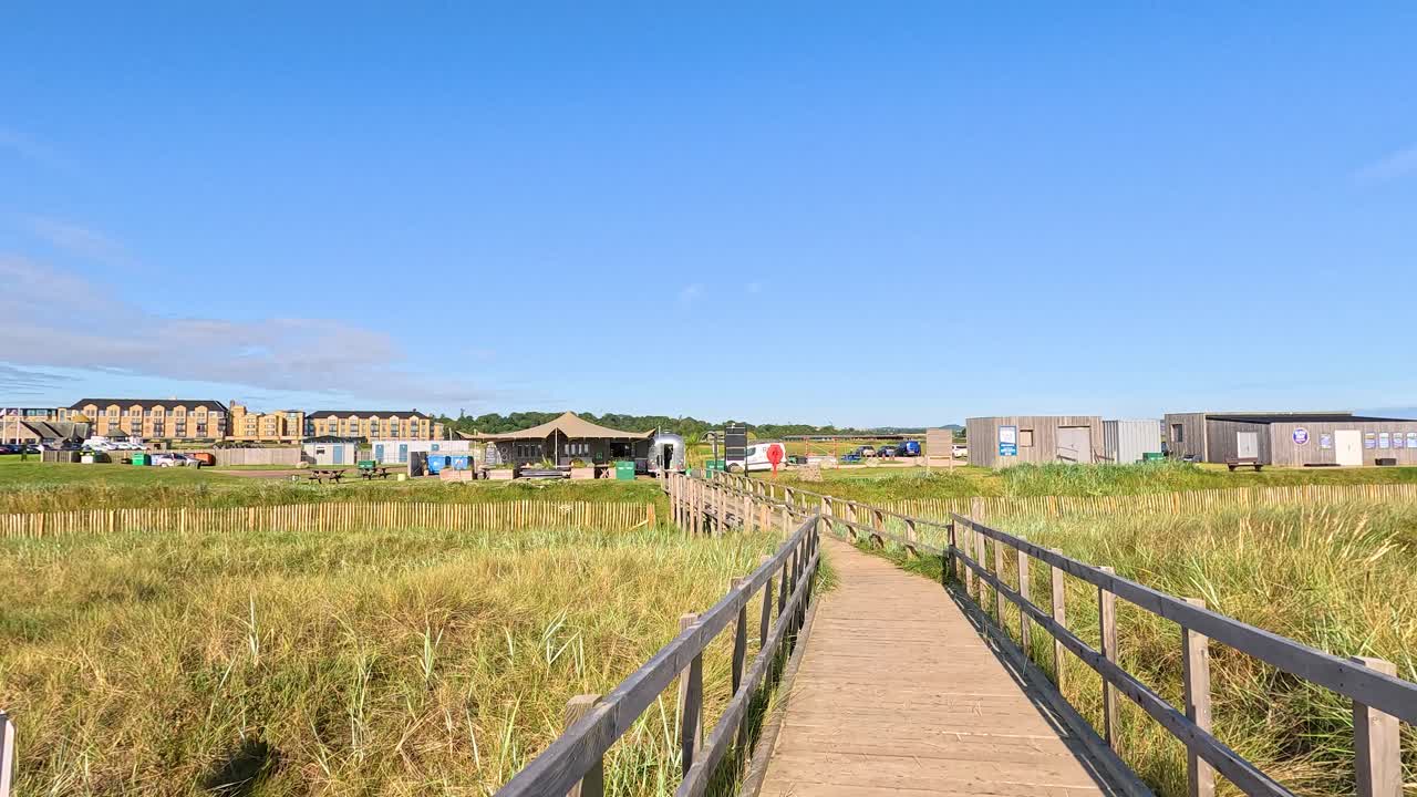 Forward motion along wooden boardwalk through grassy dunes, bright sunlight, clear blue sky, static camera