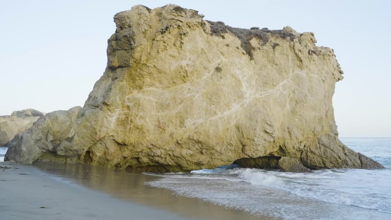 Ocean waves wash against a large rock formation at the shoreline under soft daylight on a calm, empty beach