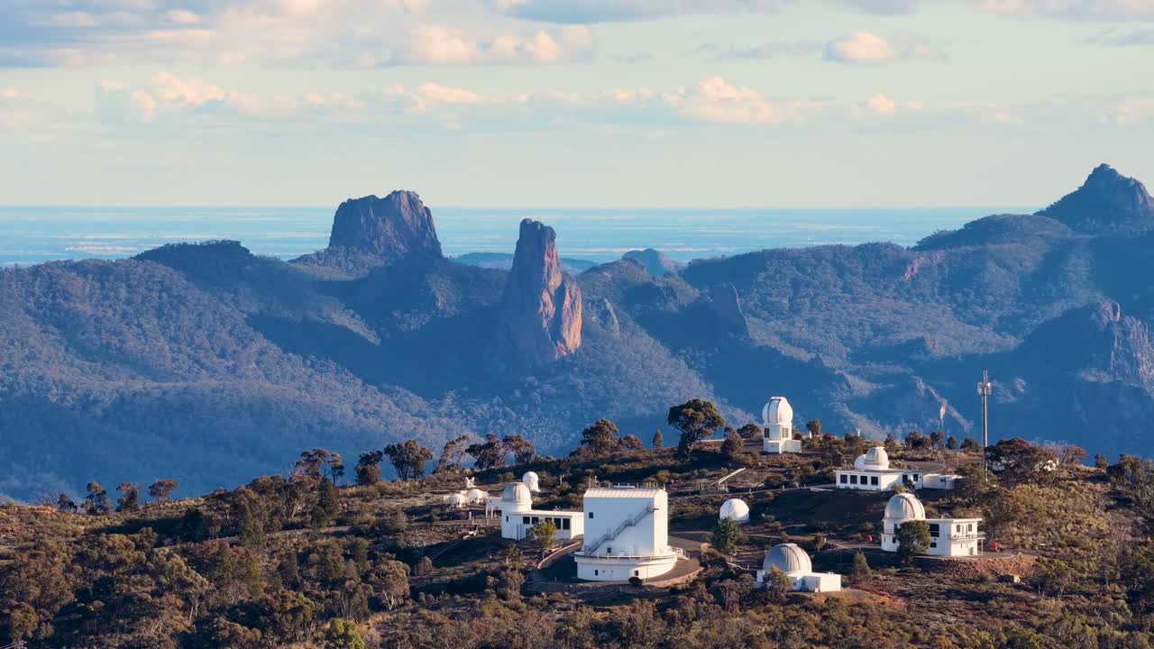 Drone camera smoothly pans over a mountaintop astronomical observatory complex with telescopes, set against rugged blue mountains and warm sunset lighting