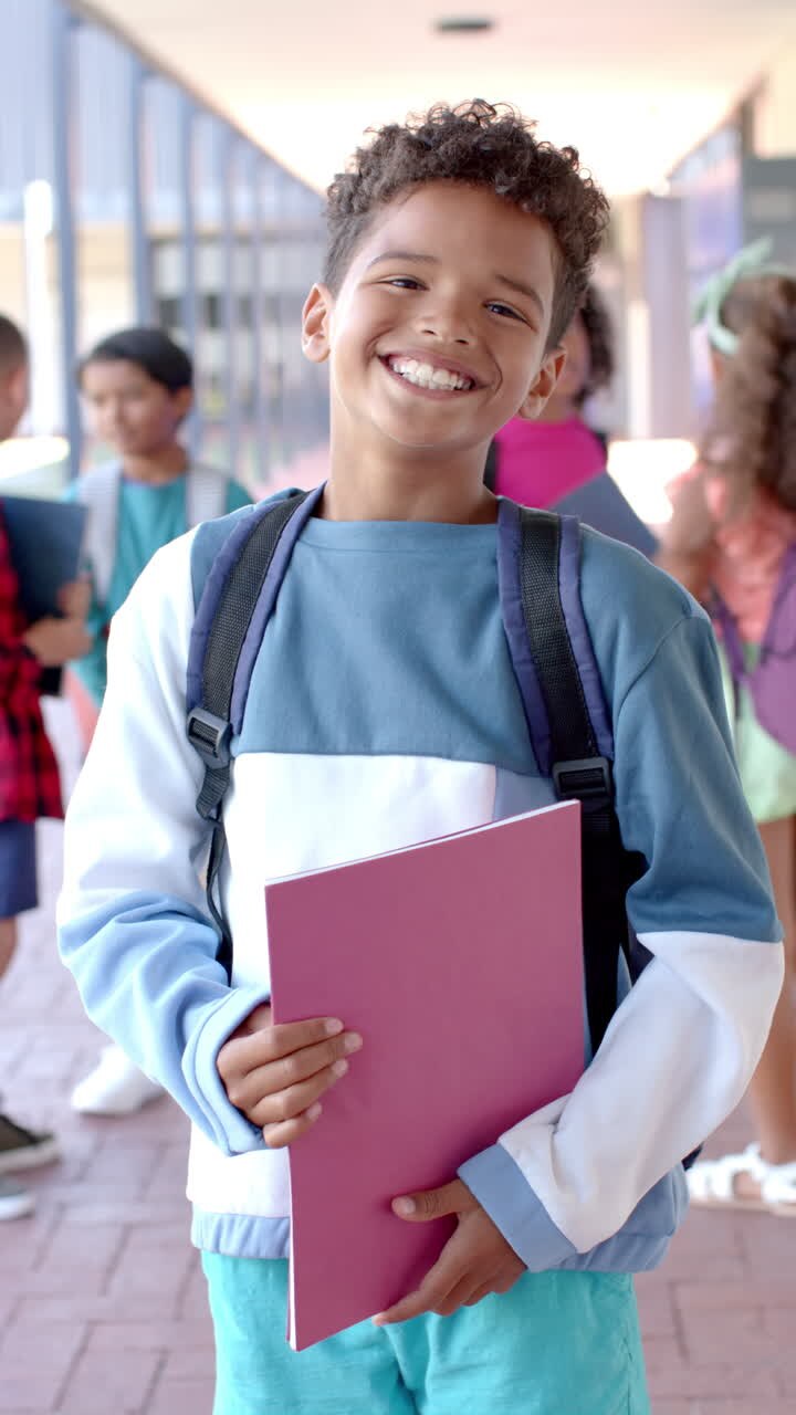 Video vertical: En la escuela, un niño pequeño sosteniendo libros y llevando una mochila está sonriendo.