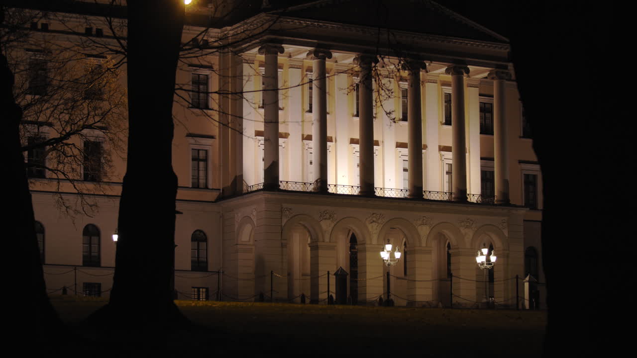 Stabilized slow motion wide 4K shot of dark trees and branches moving in parallax motion in front of illuminated Norwegian Royal Palace building facade, at night in Oslo Norway