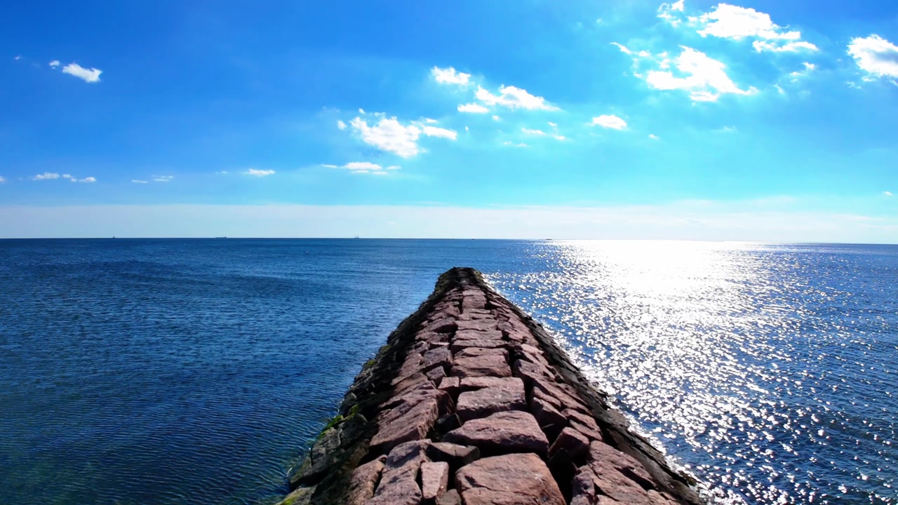 Aerial dolly view of a stone pier extending into the calm blue sea under a bright sky. A tranquil coastal scene with horizon, sunlight reflections, and peaceful ocean atmosphere